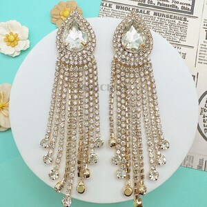 May include: A pair of gold tone dangle earrings with a teardrop shaped crystal in the center of each earring. The earrings are made of a chain of small crystals.