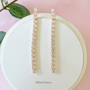 May include: A pair of gold-toned, linear drop earrings. Each earring features a row of clear, square-cut crystals set in a gold-colored metal. The earrings are displayed on a white, circular surface against a pink backdrop. The brand name "MiniChoco" is visible.