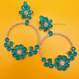 May include: A pair of large hoop earrings with a floral design. The earrings are made of silver and have green gemstones. The earrings are a statement piece and would be perfect for a night out.