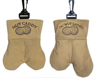 May include: Two tan-colored golf ball pouches, each with a black clip. The pouches are shaped like a pair of testicles. The text "NUT CADDY" and "OL' NUT CADDY" is printed on the front, along with a graphic of two peanuts.