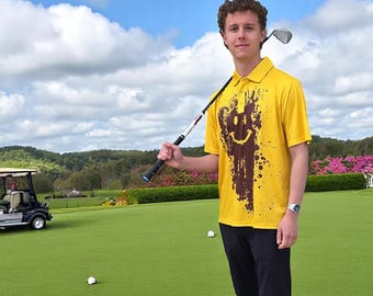 May include: A yellow polo shirt with a brown smiley face design. The shirt is worn with black pants and white sneakers. A golf club rests on the shoulder. The background shows a golf course with a cart and green grass.