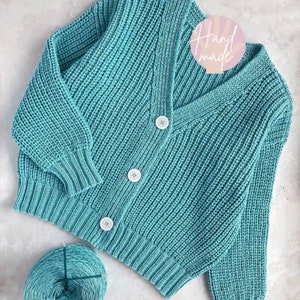 Puede incluir: Un cárdigan de punto azul turquesa con cuello en V y tres botones blancos. El cárdigan está hecho a mano y está sobre una superficie blanca con un ovillo de lana azul turquesa.