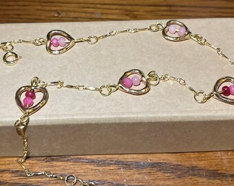 May include: A gold chain bracelet with pink heart charms. Each heart charm has two pink beads inside.
