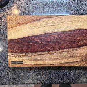 May include: Rectangular wooden cutting board with a central dark red resin river. The board features light and dark wood grain patterns. A small metal label is affixed to the lower left corner. This handcrafted board is suitable for kitchen use.