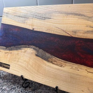 May include: A rectangular wooden cutting board with a river of red and blue resin. The wood has a light brown color with dark streaks and knots. A small metal plaque is visible on the left side.