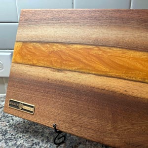 May include: A rectangular wooden cutting board with a central band of amber-colored resin. The wood has a rich, dark brown color with visible grain. A small metal plaque with text is affixed to the board. A black metal stand is attached.