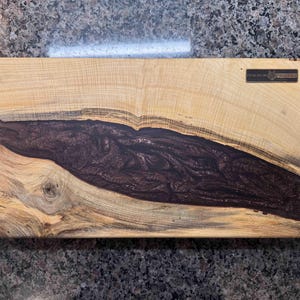 May include: Rectangular wooden cutting board with a dark brown epoxy river design. The light-colored wood grain contrasts with the dark epoxy. A small metal plaque is affixed to the top right corner.