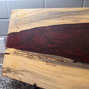 May include: Wooden cutting board with a river of deep red epoxy resin. The board is made of light-colored wood with dark gray streaks and natural wood grain. The epoxy resin creates a striking contrast.