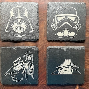 May include: Four square black slate coasters with white etched designs. Two coasters feature Darth Vader's helmet and figure, and two feature a Stormtrooper helmet. The coasters are arranged on a dark wood surface.