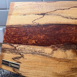 May include: A rectangular wooden cutting board with a dark reddish-brown epoxy resin river. The wood grain is light brown with dark streaks. A metal tag with the text "DESIGNS BY CHADWICK" is attached to the board.