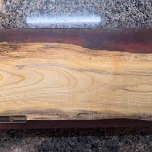May include: Rectangular wooden cutting board with a natural wood grain pattern. The board features a combination of light and dark wood tones, with a dark reddish-brown border. A small metal tag with text is affixed to the board.