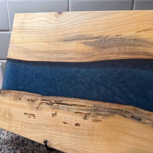 May include: Wooden cutting board with a blue epoxy resin river. The board is made of light-colored wood with dark grain patterns and natural imperfections. The blue resin creates a contrasting river effect, adding a decorative element to the kitchen item.