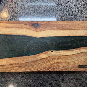 May include: A rectangular wooden serving board with a dark green epoxy river design. The wood has a natural grain pattern with varying shades of brown. A small metal plaque is affixed to the bottom right corner. This decorative board is suitable for serving food.