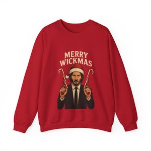 Könnte beinhalten: Ein rotes Sweatshirt mit Rundhalsausschnitt und einer Grafik eines Mannes im schwarzen Anzug mit Weihnachtsmütze, der zwei pistolenförmige Zuckerstangen hält. Darüber steht der Text MERRY WICKMAS in heller, großer Schrift.