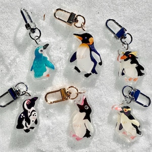 May include: A collection of acrylic penguin keychains in various colors and designs. Each keychain features a different penguin illustration, with details like blue, black, white, and yellow. The keychains have silver and gold-colored clasps.