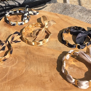 Peut inclure: Cinq bracelets circulaires composés de maillons de chaîne en métal doré entrelacés avec des rubans en tissu. Les rubans présentent divers motifs et couleurs, notamment un imprimé léopard noir et blanc, doré uni, noir uni, léopard marron et blanc uni.