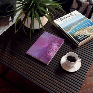 May include: A pink and purple notebook with a textured cover sits on a dark wood table with a cup of coffee and a travel magazine. The magazine cover features the text "TRAVEL MAGAZINE" and a photo of a coastal landscape.