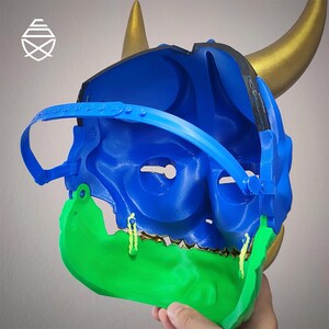 Op de afbeelding: Een blauw en groen masker met gouden hoorns. Het masker heeft een blauw interieur, een groene kaak en een blauwe band. De hoorns zijn goudkleurig. Het masker is waarschijnlijk voor cosplay of kostuums.