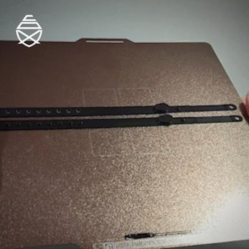 Op de afbeelding: Twee zwarte leren riemen met meerdere gaten en een gesp. De riemen liggen op een getextureerd, bronskleurig oppervlak. De riemen lijken voor een tas of ander accessoire te zijn.