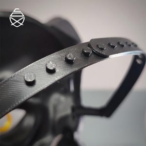 Op de afbeelding: Close-up van een zwart metalen object met een gebogen, geklonken band. De band heeft verschillende kleine, zilverkleurige klinknagels en een getextureerd oppervlak. Het object lijkt deel uit te maken van een grotere structuur, mogelijk een helm of masker.