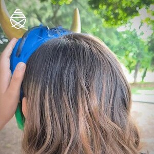 Op de afbeelding: Een blauw en goud gehoofdbescherming wordt op het hoofd van een persoon geplaatst. De hoofdbescherming heeft gouden hoorns en een blauwe stoffen bekleding. Het haar van de persoon is bruin met blonde highlights. De achtergrond is een wazig beeld van groene bomen.