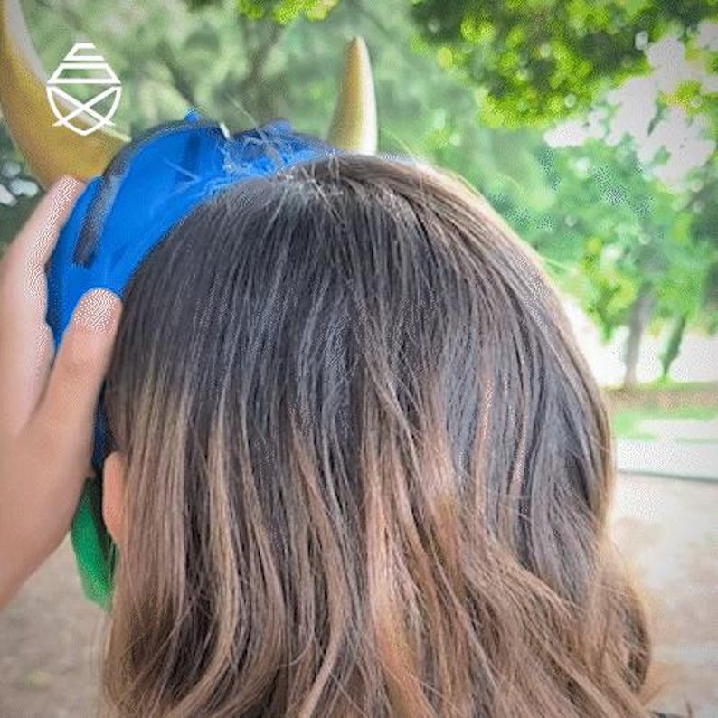Op de afbeelding: Een blauw en goud gehoofdbescherming wordt op het hoofd van een persoon geplaatst. De hoofdbescherming heeft gouden hoorns en een blauwe stoffen bekleding. Het haar van de persoon is bruin met blonde highlights. De achtergrond is een wazig beeld van groene bomen.
