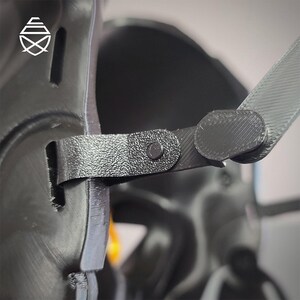 Op de afbeelding: Close-up van een zwart, getextureerd object met een riem en een klinknagel. De riem is verbonden met een rond, plat stuk. Het object heeft een geometrisch logo in de linkerbovenhoek. De achtergrond is wazig.