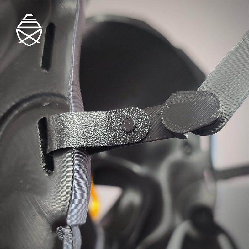 Op de afbeelding: Close-up van een zwart, getextureerd object met een riem en een klinknagel. De riem is verbonden met een rond, plat stuk. Het object heeft een geometrisch logo in de linkerbovenhoek. De achtergrond is wazig.