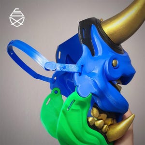 Op de afbeelding: Een blauw, groen en goud Oni-masker met een gedetailleerd ontwerp. Het masker heeft een blauw gezicht met gouden hoorns en tanden, groene accenten en een blauwe band om te dragen. Het masker is een cosplay- of decoratie-item.