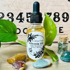 May include: A glass bottle of essential oil labeled "Occulture Crypt Artemis Oil Cedar, Patchouli, Juniper Berry" with a green plant, crystals, and a wooden board with letters and numbers in the background.