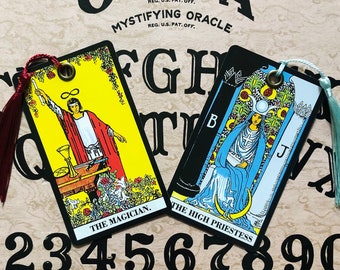 Puede incluir: Dos cartas de tarot, El Mago y La Sacerdotisa, se colocan sobre una tabla de Ouija de madera con las letras FGH y los números 1234567890 visibles. Las cartas son amarillas y negras con ilustraciones y texto coloridos.