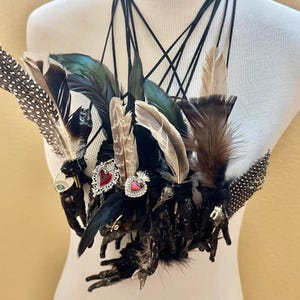May include: A striking feather necklace, handmade with a variety of feathers in brown, black, and teal. The necklace is adorned with small heart-shaped embellishments. The necklace is displayed on a mannequin.