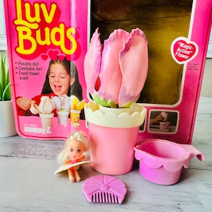 Könnte beinhalten: Ein Luv Buds Spielzeugset bestehend aus einem rosa Kunststoff-Blumentopf mit einem großen, tulpenförmigen rosa Deckel, einer kleinen blonden Puppe, einem rosa Kamm und einem rosa gewellten Becher. Die rosa Kartonverpackung zeigt den Text Luv Buds.