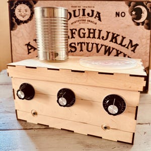 May include: A wooden box with three black knobs and a metal can on top. A clear plastic lid sits next to the can. The background features a Ouija board with the words "Ouija" and "Mystifying Oracle".