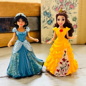 May include: Two princess figurines. One wears a blue dress with a silver bodice and a glittery skirt. The other wears a yellow dress with a red rose design on the skirt. Both have dark hair and are smiling.