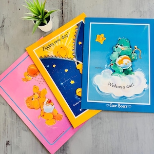 May include: Three Care Bears notebooks in pink, yellow, and blue. The pink notebook shows a Care Bear playing basketball. The yellow notebook has a sun and the text "Zippity new day!". The blue notebook has a Care Bear on a cloud with the text "Wish on a star!"