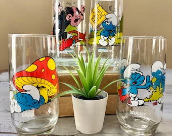 Può includere: Set di cinque bicchieri vintage dei Puffi. Ogni bicchiere presenta una diversa illustrazione del personaggio dei Puffi con etichette di testo. I bicchieri sono trasparenti con grafiche colorate e sono disposti con una piccola pianta in vaso al centro.