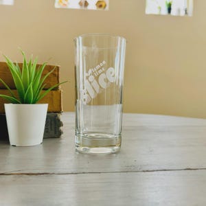 Pu&ograve; includere: Un bicchiere di vetro trasparente con le parole "lemon lime slice" in bianco, con un carattere stilizzato. Il bicchiere &egrave; cilindrico con linee verticali e poggia su una superficie grigio chiaro. Una piccola pianta in vaso e libri sullo sfondo.
