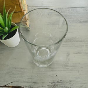 Pu&ograve; includere: Un bicchiere da bevanda trasparente con una forma leggermente svasata. Il bicchiere ha un motivo floreale vicino alla base. Una piccola pianta succulenta in vaso e un libro sono sullo sfondo, su una superficie di legno imbiancato.