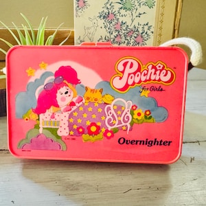 May include: A vintage pink metal lunchbox with a cartoon graphic of a dog-like character in pajamas, a cat, and the text "Poochie for Girls" and "Overnighter". The lunchbox has a white handle.