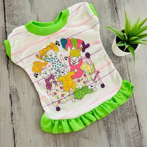 May include: White pet shirt with a green collar and ruffled hem. The shirt features a colorful graphic of cartoon cats with the word "PARTY" above them. The shirt has horizontal stripes in various colors.