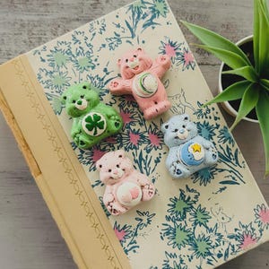 Puede incluir: Cuatro figuras de osos cariñosos de colores sobre un libro vintage con estampados florales. Los osos son rosas, verdes y azules, cada uno con insignias únicas en el vientre. El libro tiene una cubierta color crema y un lomo decorativo. Una pequeña planta en maceta está en el fondo.