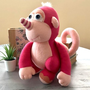 Op de afbeelding: Een roze en lichtroze pluche aap. Het speelgoed heeft een gestreepte neus, grote blauwe ogen en een gekrulde staart. De aap zit op een licht oppervlak, met een kleine plant in een pot en een boek op de achtergrond.