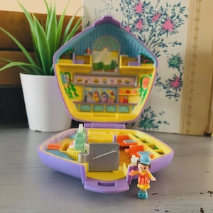 May include: A vintage Polly Pocket playset, open to reveal a miniature diner scene. The playset is purple, yellow, and blue, with a small figure of a person standing in front. The diner has a counter with food and seating areas.