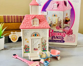 Può includere: Un playset Fisher-Price Precious Places con una casa con tetto rosa e facciata bianca, una chiave, una carrozza trainata da cavalli e una scatola. La casa ha un cartello con scritto "Precious Places". La scatola dice "Ages 5 and up".