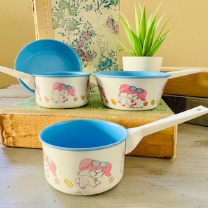 May include: A set of vintage children's cookware featuring a white enamel finish with blue interiors and handles. Each piece is decorated with a cartoon dog wearing sunglasses and a pink bow. The set includes a saucepan, a bowl, and a lid.