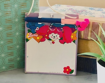 May include: A vintage Poochie notepad with a cartoon character design. The notepad has a white writing surface, a pink and purple border, and a decorative flower. It has a blue and white handle and a pink clip.