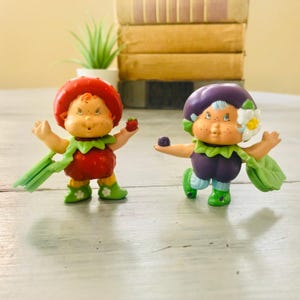 Pu&ograve; includere: Due statuette vintage di Fragolina. Una &egrave; rossa e l'altra viola. Entrambe hanno lentiggini, dettagli verdi e tengono piccoli frutti. Quella rossa tiene una fragola, e quella viola tiene un piccolo frutto rotondo, viola scuro.