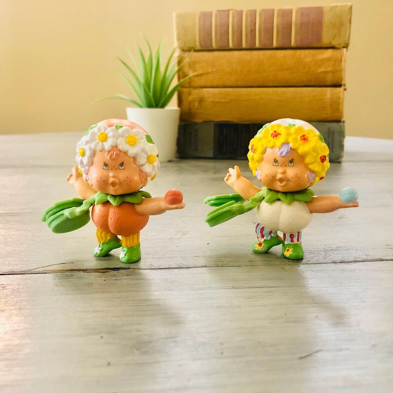 Pu&ograve; includere: Due figurine da collezione vintage. Una &egrave; arancione con una corona di margherite e l'altra &egrave; bianca con una corona di fiori gialli. Entrambe hanno sciarpe verdi a forma di foglia e tengono piccole palline. Le figurine sono su una superficie di legno chiaro.