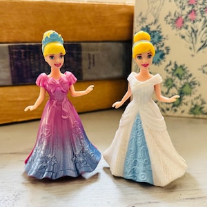 May include: Two Cinderella dolls. One doll wears a pink and blue ombre gown with a bow. The other doll wears a white gown with a blue underskirt. Both dolls have blonde hair and blue headbands.
